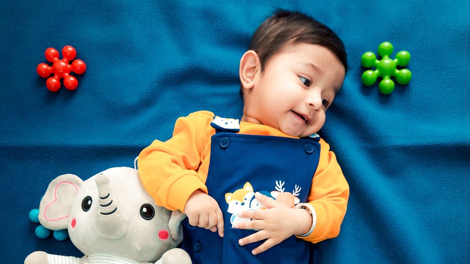 A cute baby boy lying on a vibrant blue blanket with a stuffed elephant toy and colorful toys.
