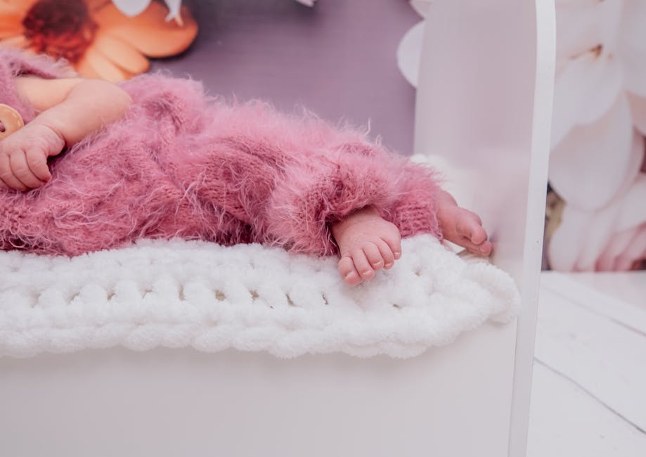 Cute baby in a pink fluffy outfit resting on a white knitted blanket with floral backdrop.